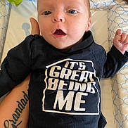 Stanley is registered to the contest to win money with this photo: baby, infant, child, person, face, clothing, shirt, tattoo, arm, hand, bed, pillow, cushion, text_on_clothing, blue, black, white, expression, surprised, indoor
