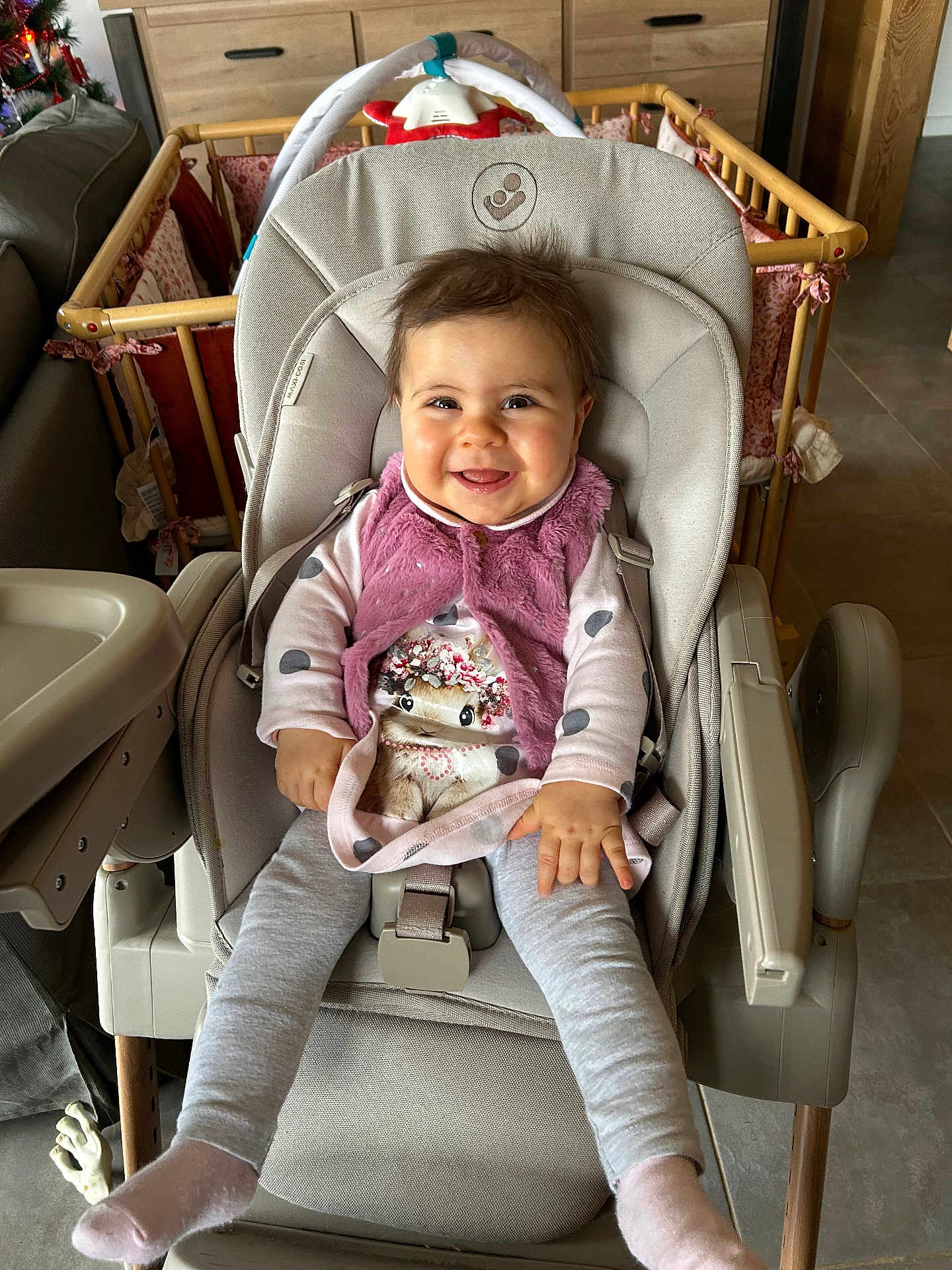 Pauline participe au concours pour gagner de l'argent avec cette photo : baby, high_chair, smiling, indoor, child, pink_vest, polka_dot, animal_print, playpen, wooden_furniture, cozy, happy, toddler, seatbelt, floor_tile, long_sleeves, socks, hair, face, person