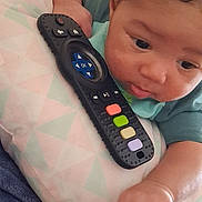 Aurelio is registered to the contest to win money with this photo: baby, infant, remote_control, pillow, curious, hand, face, eyes, clothing, soft_fabric, indoor, closeup, child, lying_down, colorful_buttons, skin, couch, relaxing, young_child, exploring