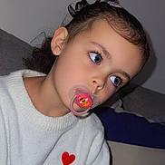 Giulia participe au concours pour gagner de l'argent avec cette photo : child, pacifier, white_sweater, red_heart, indoor, portrait, close_up, thoughtful, cute, black_hair, curly_hair, blue_eyes, face, couch, pillow, cozy, casual_clothing, kid, young_child, expression