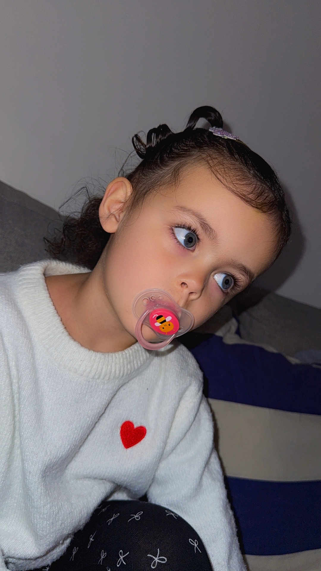 Giulia participe au concours pour gagner de l'argent avec cette photo : child, pacifier, white_sweater, red_heart, indoor, portrait, close_up, thoughtful, cute, black_hair, curly_hair, blue_eyes, face, couch, pillow, cozy, casual_clothing, kid, young_child, expression