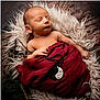 newborn, baby, sleeping, wrapped, blanket, fur, red_cloth, pendant, hand, face, peaceful, portrait, cozy, resting, infant, soft, closeup, cute, child, indoors