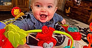 Mallory joined the competition — help win amazing prizes! activity_center, baby, blue_clothing, child, colorful, face, furniture, hand, happy, home, indoor, infant, living_room, person, plastic_toy, play, smiling, soft_toy, toy, wooden_cabinet