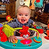 activity_center, baby, blue_clothing, child, colorful, face, furniture, hand, happy, home, indoor, infant, living_room, person, plastic_toy, play, smiling, soft_toy, toy, wooden_cabinet