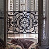 cat, sleeping, pillow, window, open_window, shutters, wrought_iron, decorative_ironwork, gray_cat, indoor, cozy, resting, natural_light, curtain, fabric, butterfly_pattern, plaid_blanket, quiet, peaceful, home