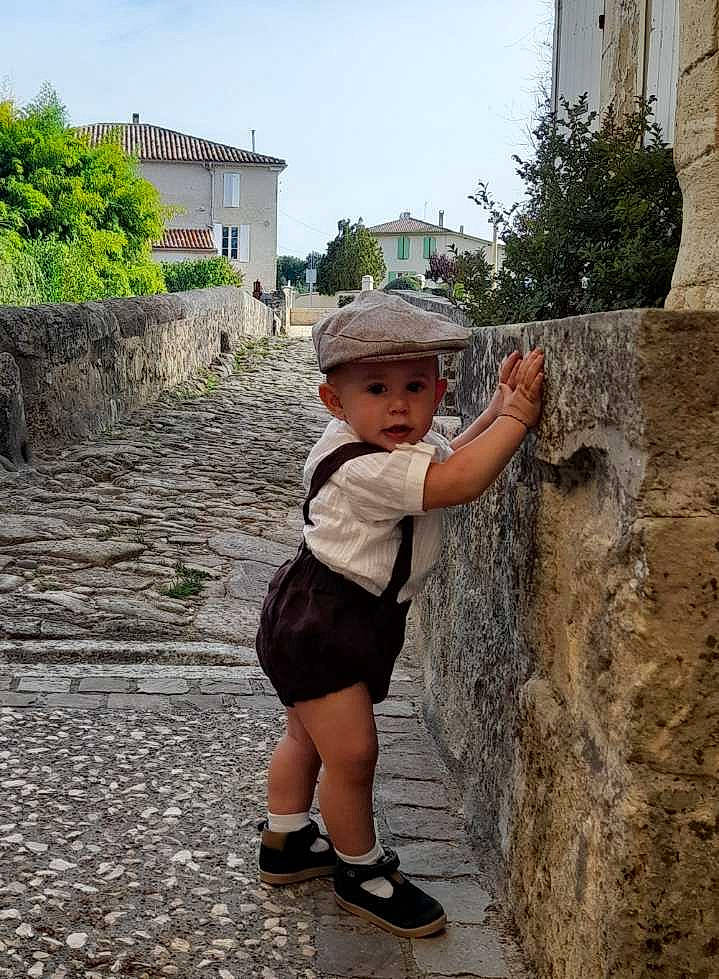 Maé a rejoint le concours — aidez-le/la à gagner de superbes lots ! bedrock, cobblestone, concrete, hat, headwear, human_leg, landscape, leg, leisure, person, plant, public_space, recreation, road, sandal, shorts, sky, summer, temple, toddler