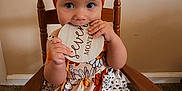 Evelyn joined the competition — help win amazing prizes! baby, child, infant, girl, orange_bow, wooden_chair, floral_dress, carpet, wall, milestone_sign, cute, portrait, indoor, smiling, hands, face, sitting, headwear, decorative, young