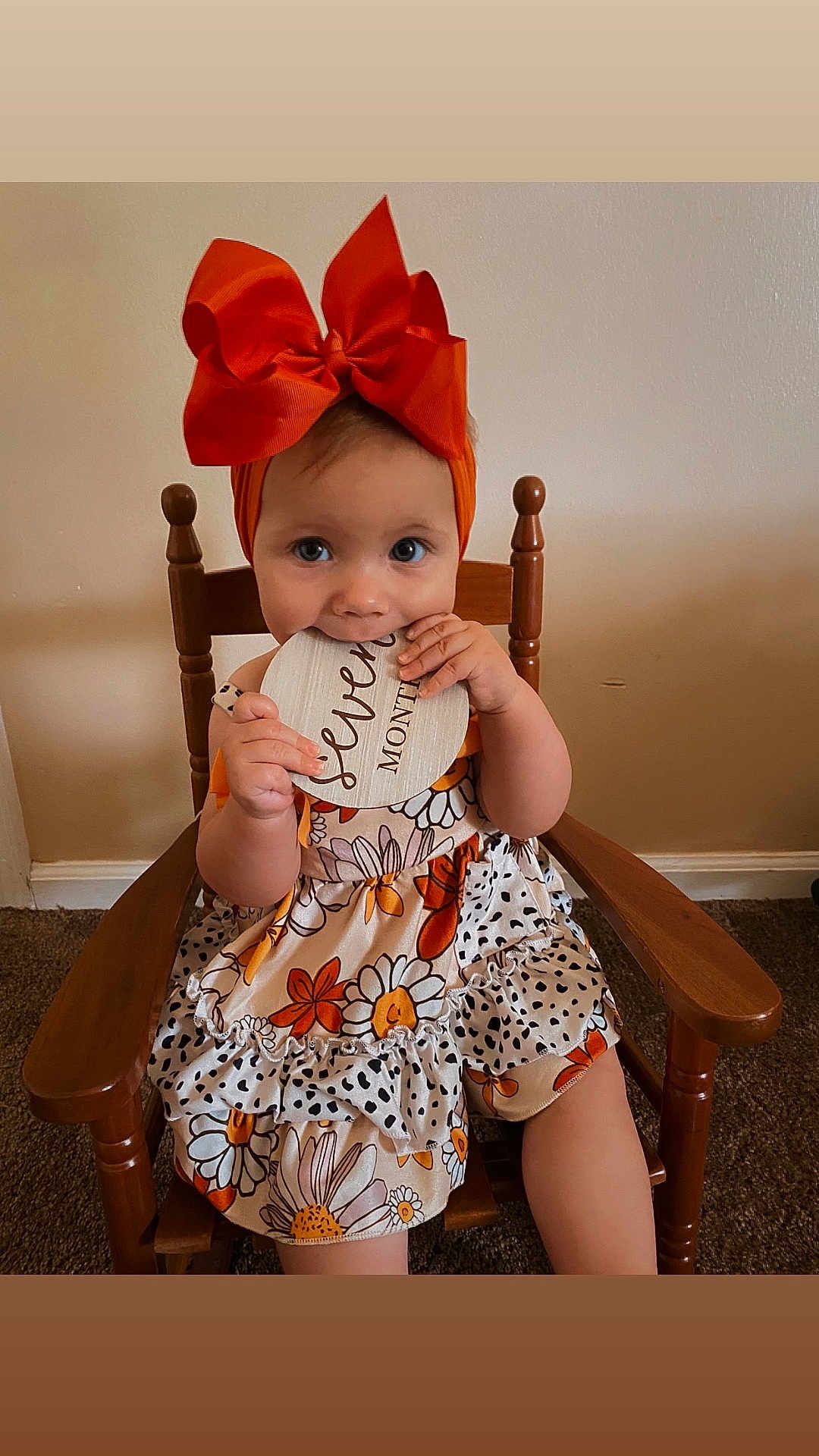 Evelyn joined the competition — help win amazing prizes! baby, child, infant, girl, orange_bow, wooden_chair, floral_dress, carpet, wall, milestone_sign, cute, portrait, indoor, smiling, hands, face, sitting, headwear, decorative, young