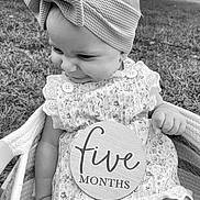 Evelyn is registered to the contest to win money with this photo: baby, infant, child, headband, bow, dress, floral_pattern, basket, grass, outdoor, black_and_white, plaque, smiling, cute, portrait, sitting, hand, face, person, young