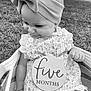 baby, infant, child, headband, bow, dress, floral_pattern, basket, grass, outdoor, black_and_white, plaque, smiling, cute, portrait, sitting, hand, face, person, young