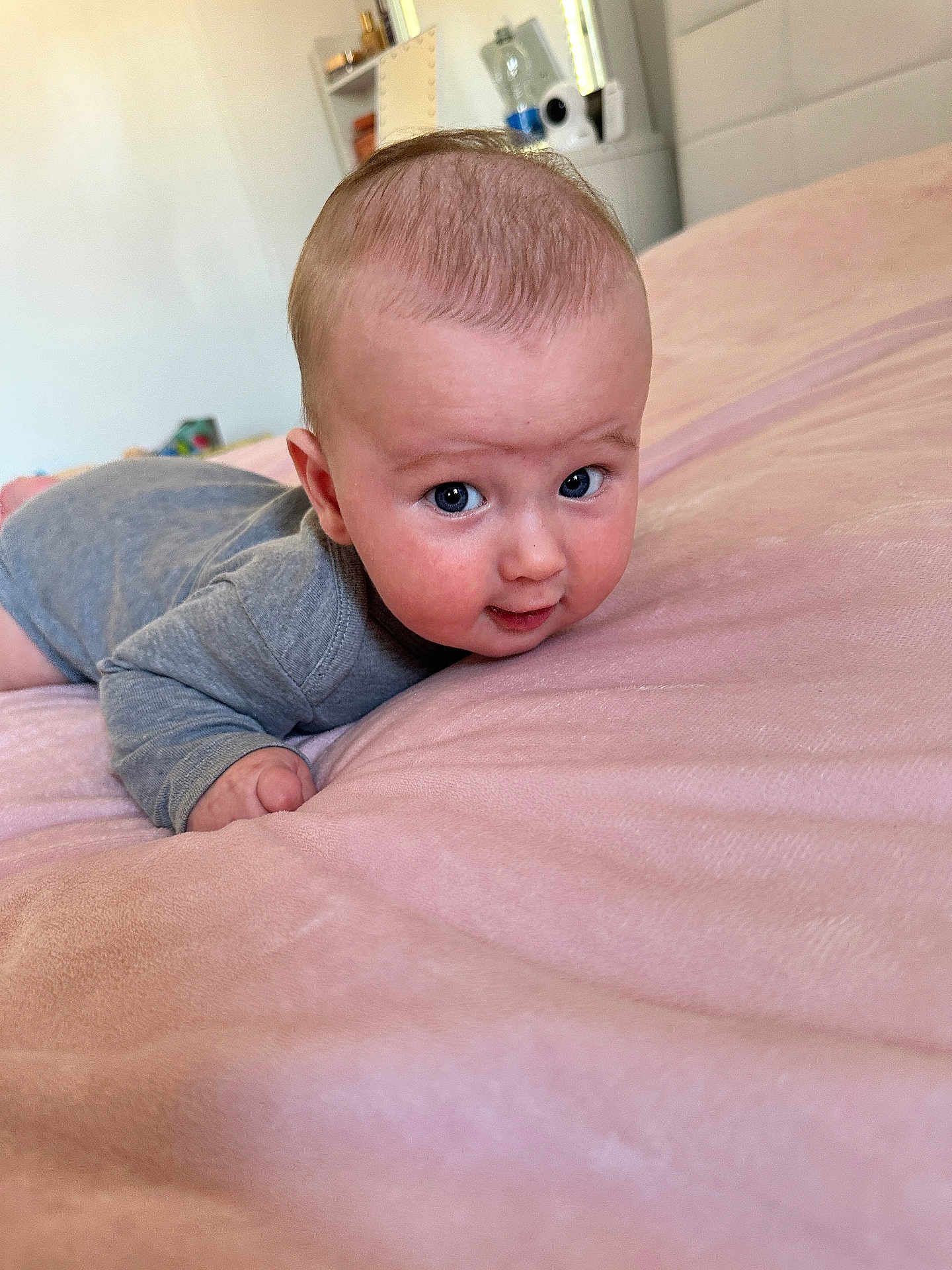 Mathias is registered to the contest to win money with this photo: baby, bedroom, child, cozy, cute, expression, eyes, face, gray_onesie, hand, head, indoors, infant, lying_down, person, pink_blanket, skin, smile, soft_texture, young_child