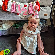 Romani joined the competition — help win amazing prizes! baby, child, party_hat, dress, carpet, crib, blanket, toy, tassel, banner, floor, indoor, decoration, cute, infant, sitting, curious, celebration, pink, white