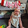 baby, child, party_hat, dress, carpet, crib, blanket, toy, tassel, banner, floor, indoor, decoration, cute, infant, sitting, curious, celebration, pink, white