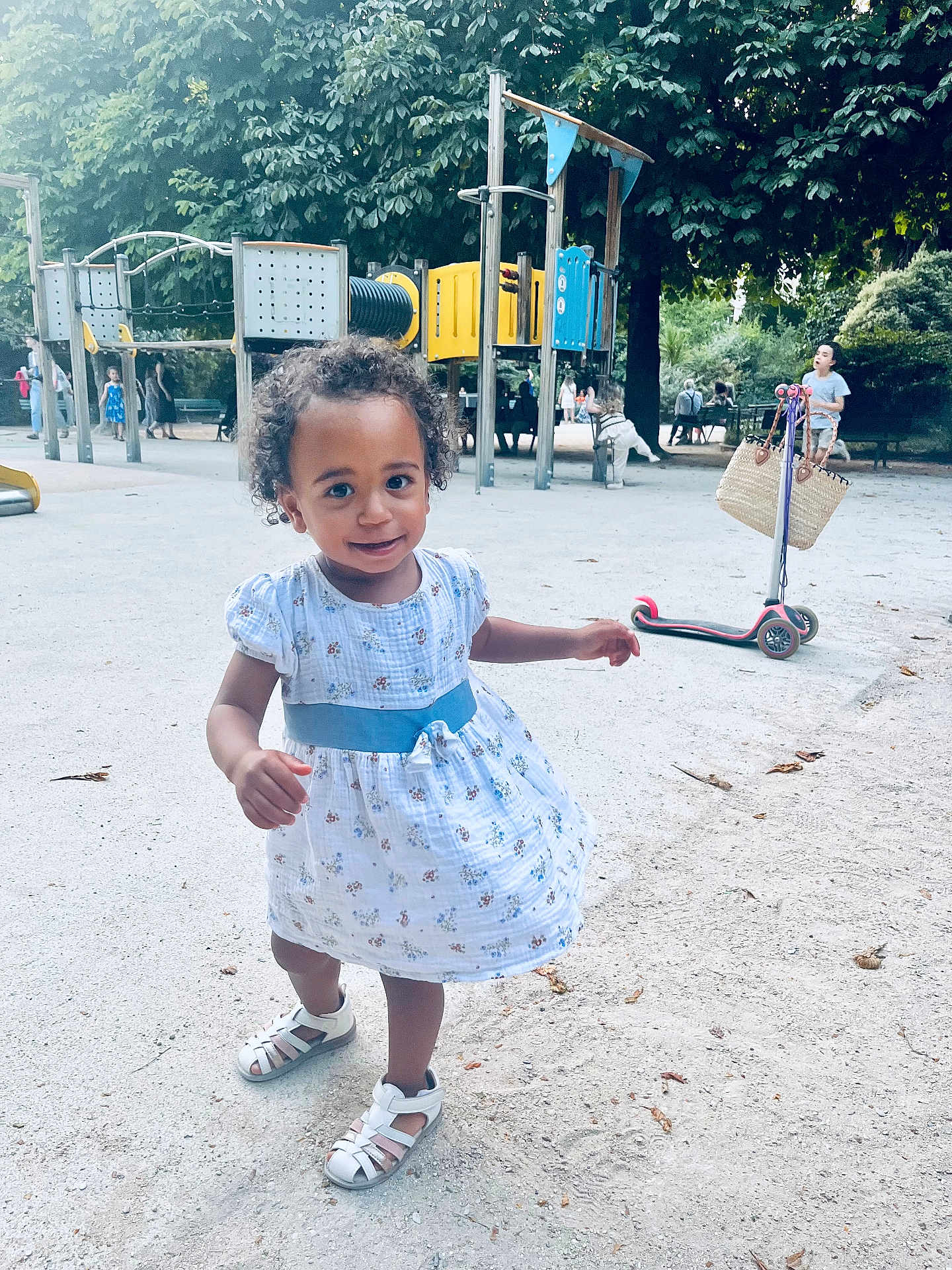 Ily a rejoint le concours — aidez-le/la à gagner de superbes lots ! basket, child, curly_hair, daylight, dress, happy, outdoor, park, person, play_equipment, playground, recreation, sand, sandal, scooter, smiling, toddler, trees, walking, young_child