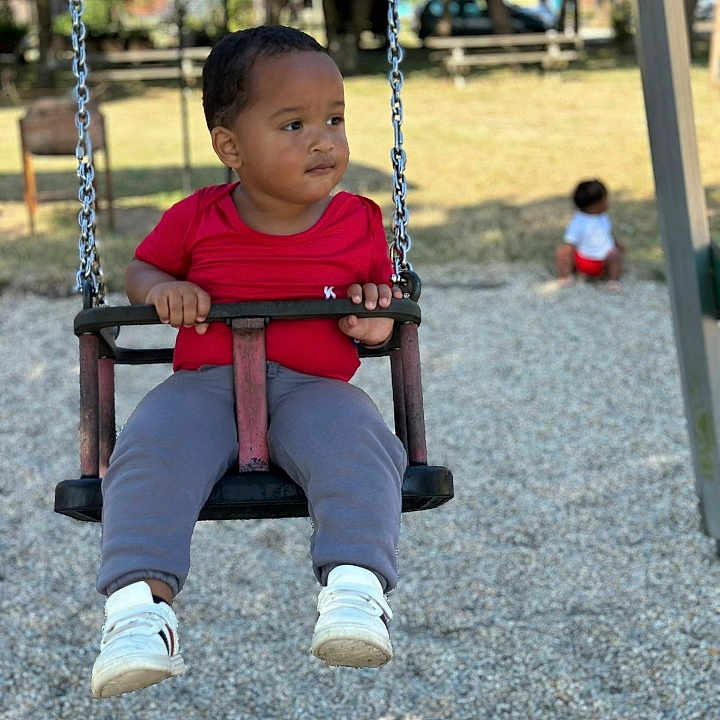 Noah -Lamine a rejoint le concours — aidez-le/la à gagner de superbes lots ! baby, background, bench, chain, child, daylight, fun, grass, gray_pants, outdoor, park, person, play, playground, red_shirt, seat, swing, trees, white_shoes, young
