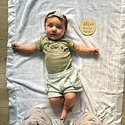 Adleigh joined the competition — help win amazing prizes! baby, infant, blanket, headband, flower, green_shirt, shorts, wooden_sign, text, floor, legs, floral_pants, toddler, lying_down, indoors, cute, child, person, soft_texture, home