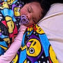 baby, sleeping, blanket, pacifier, emoji_pattern, colorful, infant, cozy, soft_lighting, closeup, face, curly_hair, pink_clothing, resting, peaceful, child, indoors, comfort, cute, young