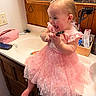 bathroom, cabinet, child, clothing, countertop, cute, hair_bow, happy, indoor, joyful, mirror, person, pink_dress, reflection, remote_control, sink, smiling, toddler, toiletries, wood_paneling