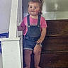 Lilia is registered to the contest to win money with this photo: toddler, child, stairs, wooden, banister, pink_shirt, denim_overalls, barefoot, indoor, curious, young_child, person, hair_tied, casual_clothing, home, wood, sitting, small_person, portrait, staircase