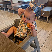 Zayn is registered to the contest to win money with this photo: toddler, child, high_chair, restaurant, crown, paper_crown, food, nugget, curious, sitting, table, floor, tile_floor, shirt, patterned_shirt, indoor, furniture, window, daylight, casual