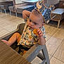 toddler, child, high_chair, restaurant, crown, paper_crown, food, nugget, curious, sitting, table, floor, tile_floor, shirt, patterned_shirt, indoor, furniture, window, daylight, casual