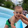 baby, child, swimming_pool, water, green_shorts, smiling, outdoor, summer, playful, person, skin, nature, trees, clouds, grass, inflatable_pool, hand, fun, happy, daytime