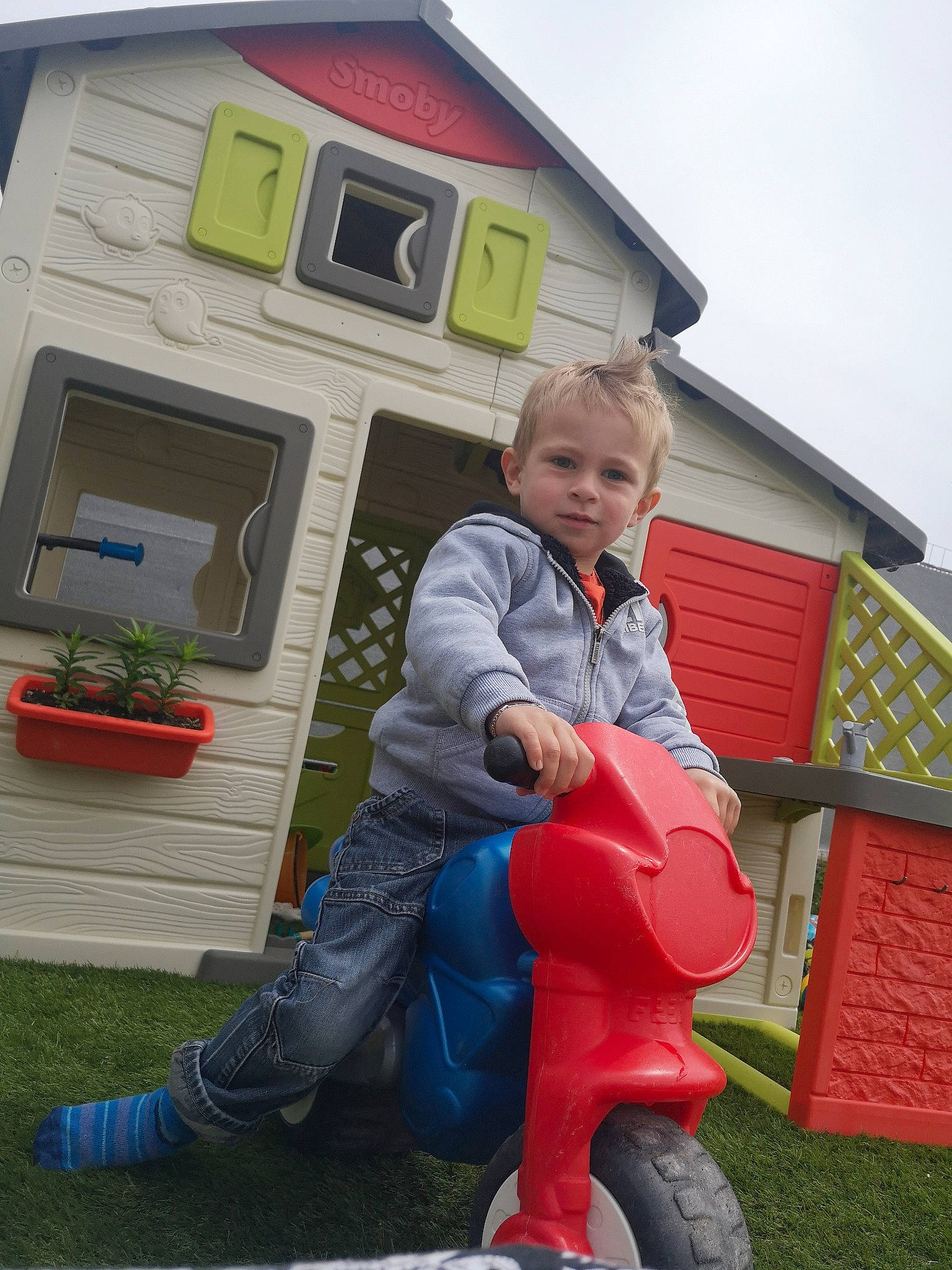 Aaron a rejoint le concours — aidez-le/la à gagner de superbes lots ! building, cottage, electric_blue, flowerpot, fun, grass, house, houseplant, jeans, lap, leisure, person, plant, recreation, riding_toy, siding, sitting, sky, tire, toddler