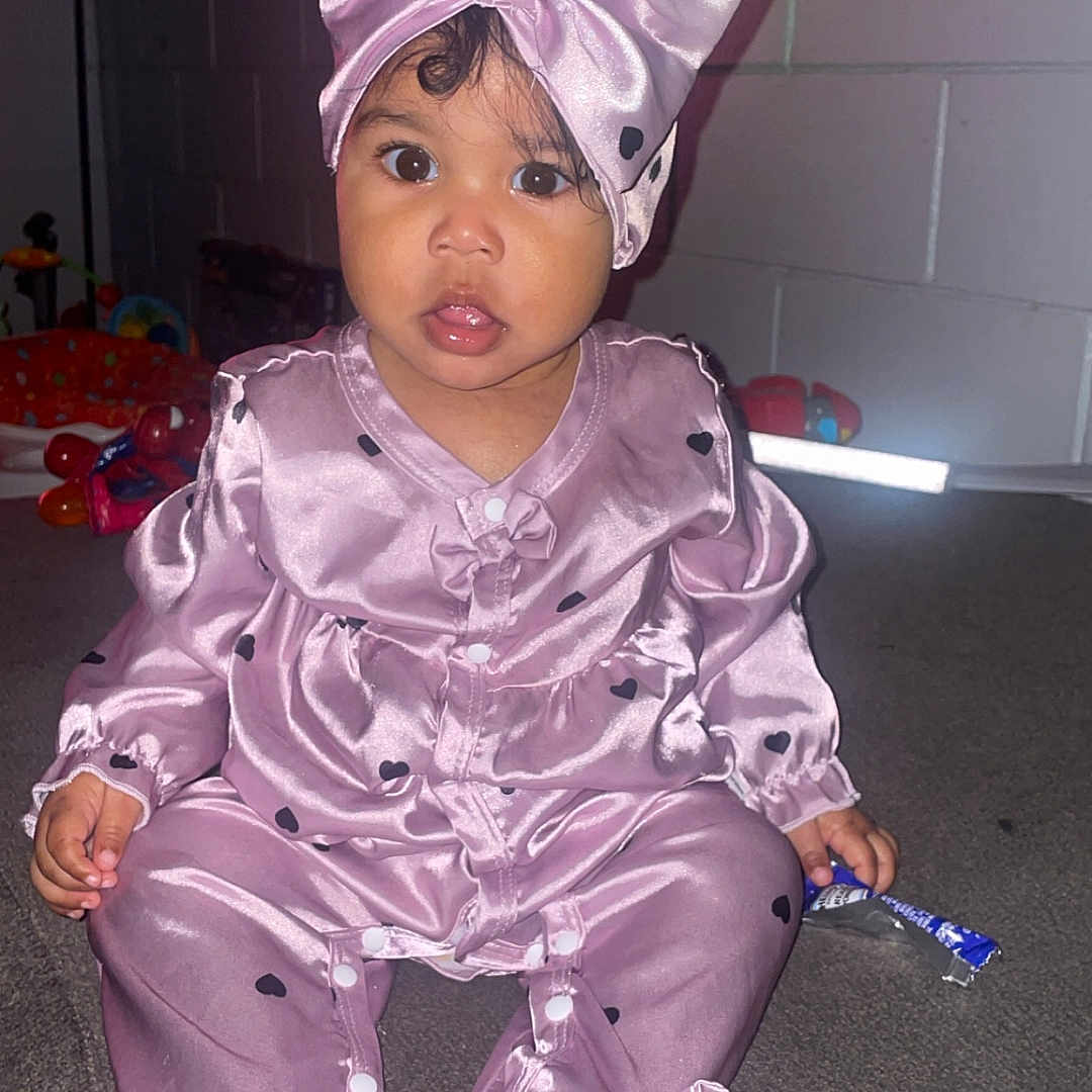 Kania is registered to the contest to win money with this photo: baby, bow, brick_wall, carpet, child, clothing, curious, cute, footed_pajamas, headband, indoor, infant, long_sleeves, person, pink_outfit, portrait, sitting, snack_wrapper, wide_eyes, young_child