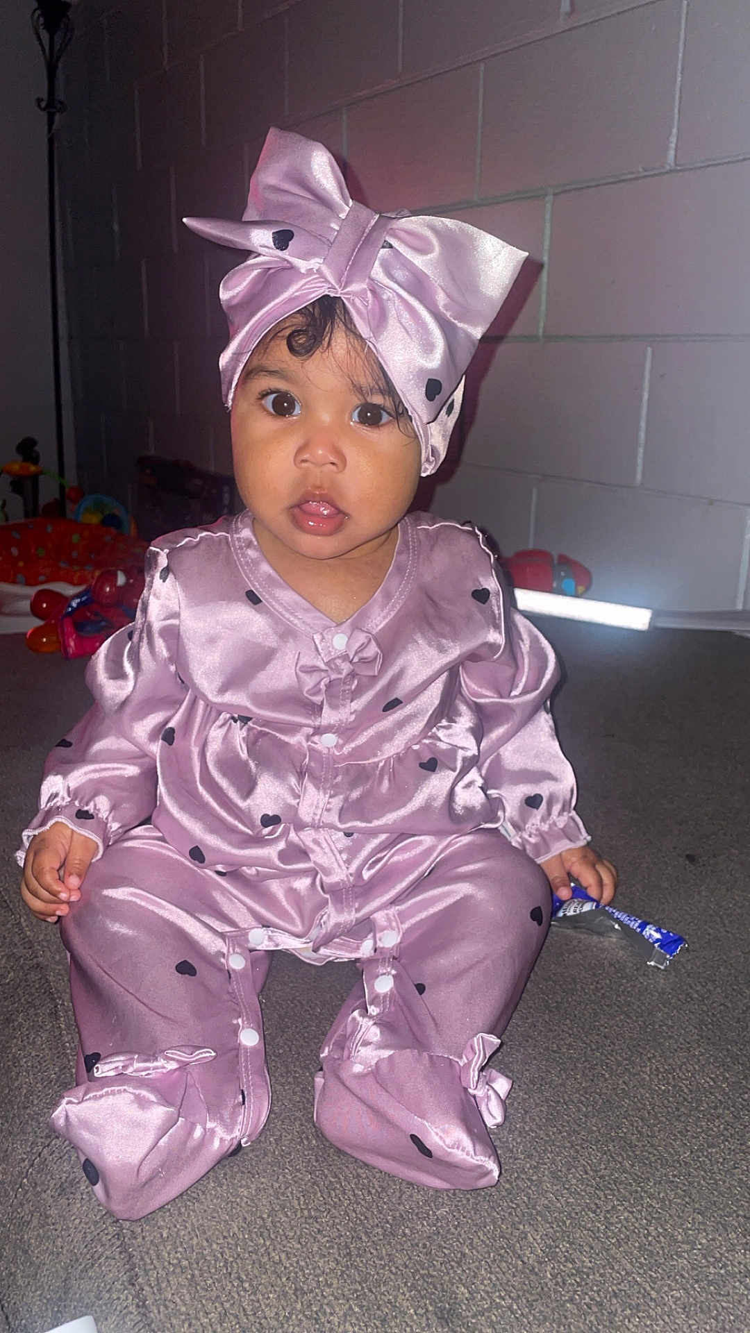 Kania is registered to the contest to win money with this photo: baby, child, pink_outfit, headband, bow, carpet, indoor, curious, wide_eyes, snack_wrapper, sitting, brick_wall, long_sleeves, footed_pajamas, infant, portrait, cute, clothing, person, young_child