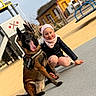 child, dog, belgian_malinois, service_dog, smiling, outdoor, playground, park, sitting, crouching, pink_hat, scarf, winter_clothing, harness, tongue_out, pointed_ears, portrait, colorful_building, sunny, play_equipment