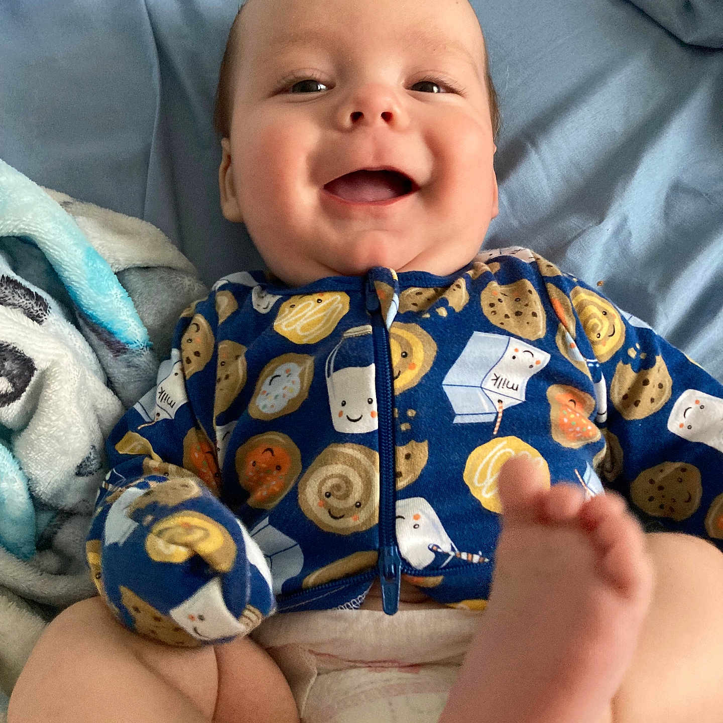 James joined the competition — help win amazing prizes! baby, smiling, onesie, cookie_pattern, milk_carton_pattern, foot, blanket, blue_background, diaper, cute, happy, infant, child, lying_down, face, skin, person, indoors, playful, closeup