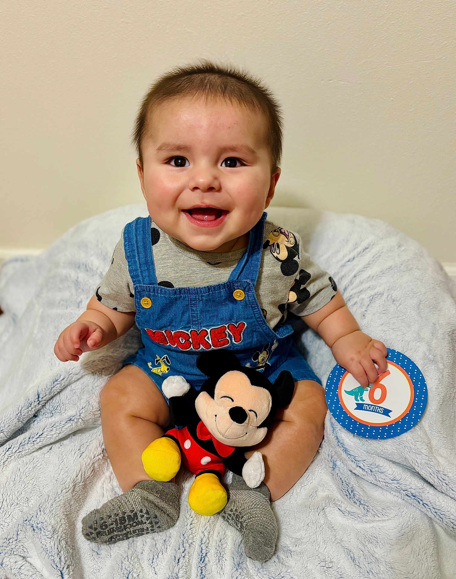 Atlas is registered to the contest to win money with this photo: baby, smiling, mickey_mouse, plush_toy, blanket, clothing, socks, milestone_card, indoor, child, happy, cute, portrait, seated, face, infant, toy, soft_texture, casual, playful