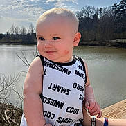Jesse is registered to the contest to win money with this photo: arm, baby, blond_hair, child, clouds, cute, face, hand, lake, outdoors, portrait, sky, sleeveless_shirt, smile, text_on_clothing, toddler, trees, water, wooden_deck, wristband