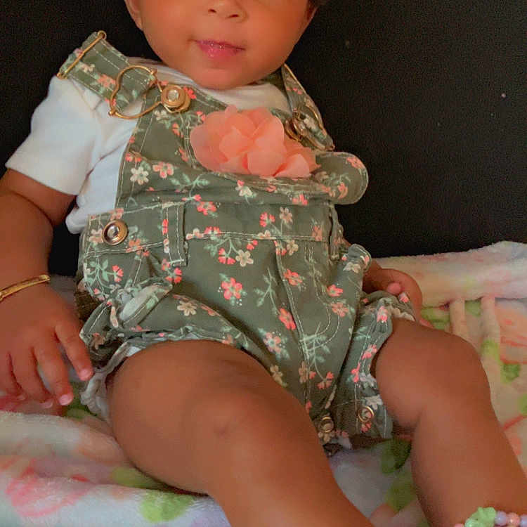 Legacy Amor is registered to the contest to win money with this photo: ankle_accessory, baby, blanket, bow, bracelet, child, clothing, colorful, cute, floral_overalls, happy, indoors, infant, person, portrait, sitting, smiling, soft_lighting, white_shirt, young_child