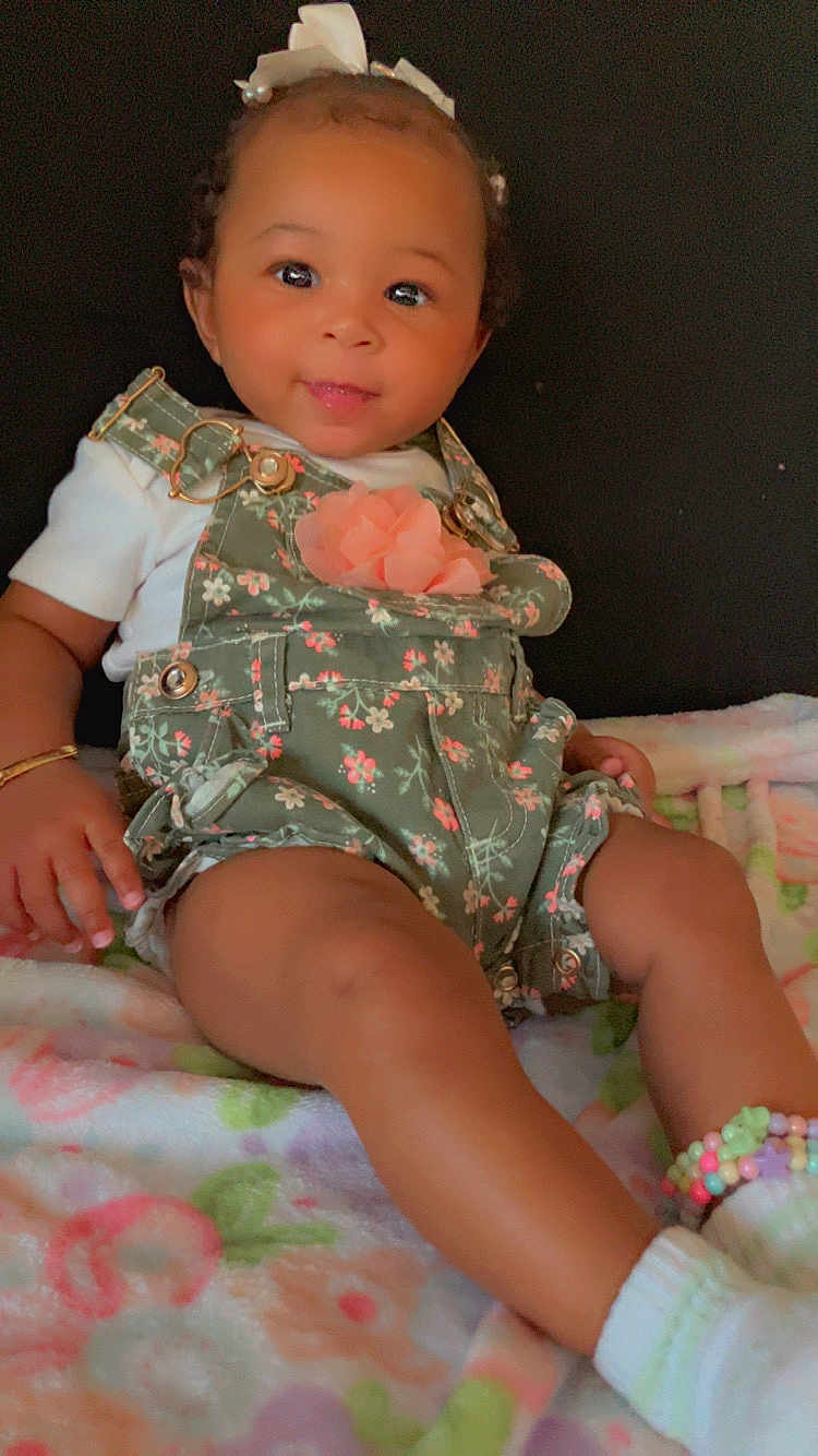 Legacy Amor is registered to the contest to win money with this photo: baby, child, infant, smiling, sitting, floral_overalls, white_shirt, bow, bracelet, blanket, colorful, cute, ankle_accessory, portrait, indoors, soft_lighting, happy, young_child, person, clothing