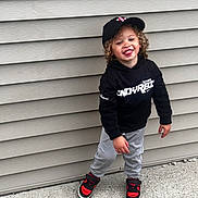 Trey joined the competition — help win amazing prizes! beige_wall, black_cap, black_hoodie, casual_pose, child, concrete, curly_hair, grey_pants, happy, headwear, outdoor, person, playful, red_sneakers, sidewalk, smiling, standing, toddler, urban, young_child
