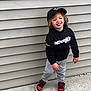 beige_wall, black_cap, black_hoodie, casual_pose, child, concrete, curly_hair, grey_pants, happy, headwear, outdoor, person, playful, red_sneakers, sidewalk, smiling, standing, toddler, urban, young_child