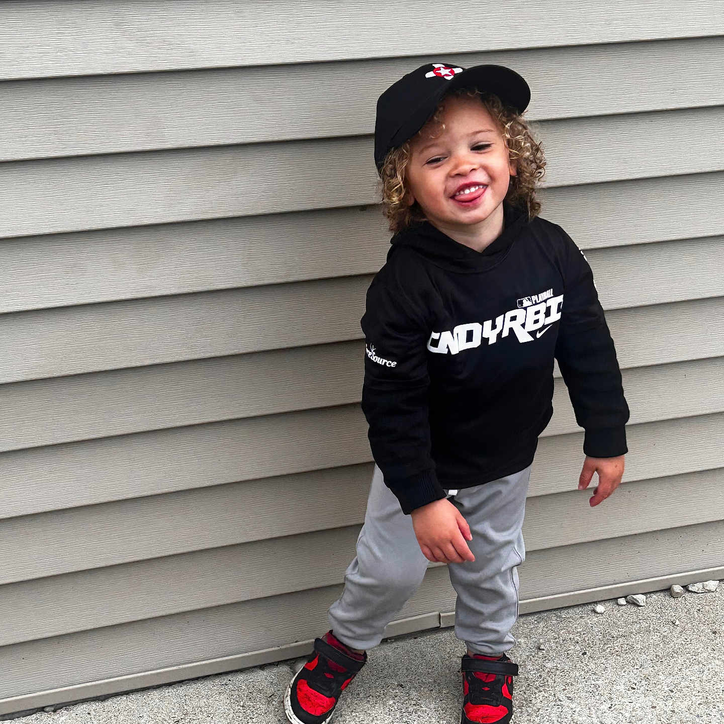 Trey joined the competition — help win amazing prizes! beige_wall, black_cap, black_hoodie, casual_pose, child, concrete, curly_hair, grey_pants, happy, headwear, outdoor, person, playful, red_sneakers, sidewalk, smiling, standing, toddler, urban, young_child