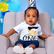 Jayden is registered to the contest to win money with this photo: baby, child, crown, half_birthday, chair, white_shirt, navy_shorts, baby_shoes, surprised_expression, indoor, portrait, celebration, balloons, soft_lighting, cute, sitting, face, headband, infant, studio_photo