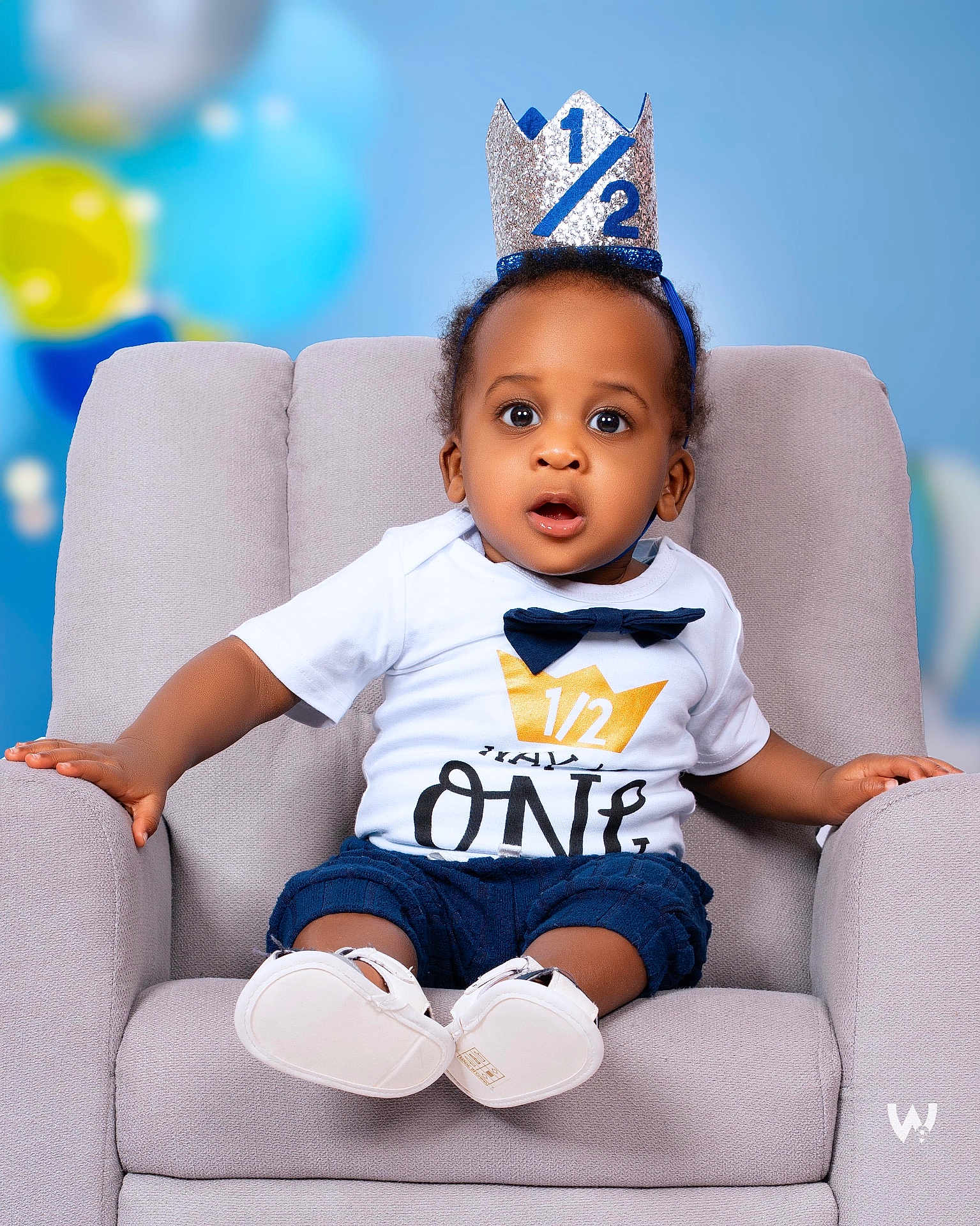 Jayden is registered to the contest to win money with this photo: baby, child, crown, half_birthday, chair, white_shirt, navy_shorts, baby_shoes, surprised_expression, indoor, portrait, celebration, balloons, soft_lighting, cute, sitting, face, headband, infant, studio_photo