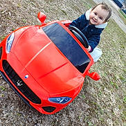 Giulya participe au concours pour gagner de l'argent avec cette photo : casual_clothing, child, cute, daytime, fun, grass, gravel, happy, jacket, outdoor, parked, person, play, red_car, small_car, smiling, steering_wheel, toddler, toy_car, vehicle