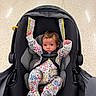 baby, infant, child, car_seat, floral_outfit, onesie, sitting, lying_down, arms_raised, blue_eyes, brown_hair, face, portrait, indoor, floor, safety_harness, travel, vehicle, parent_hand, colorful_clothing