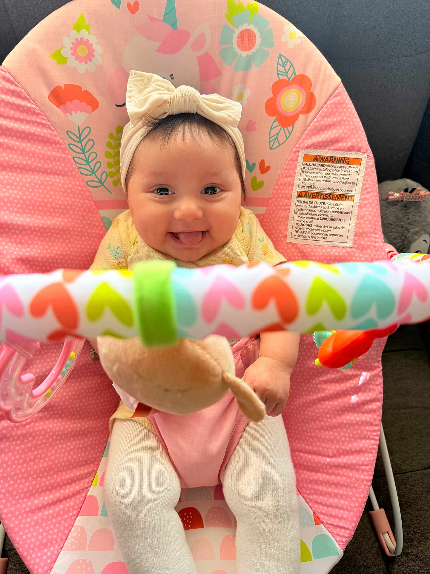 Mia is registered to the contest to win money with this photo: baby, infant, child, smiling, tongue_out, headband, bow, pink, baby_bouncer, toy_bar, colorful, hearts, flowers, plush_toy, cute, happy, sitting, indoors, closeup, playful