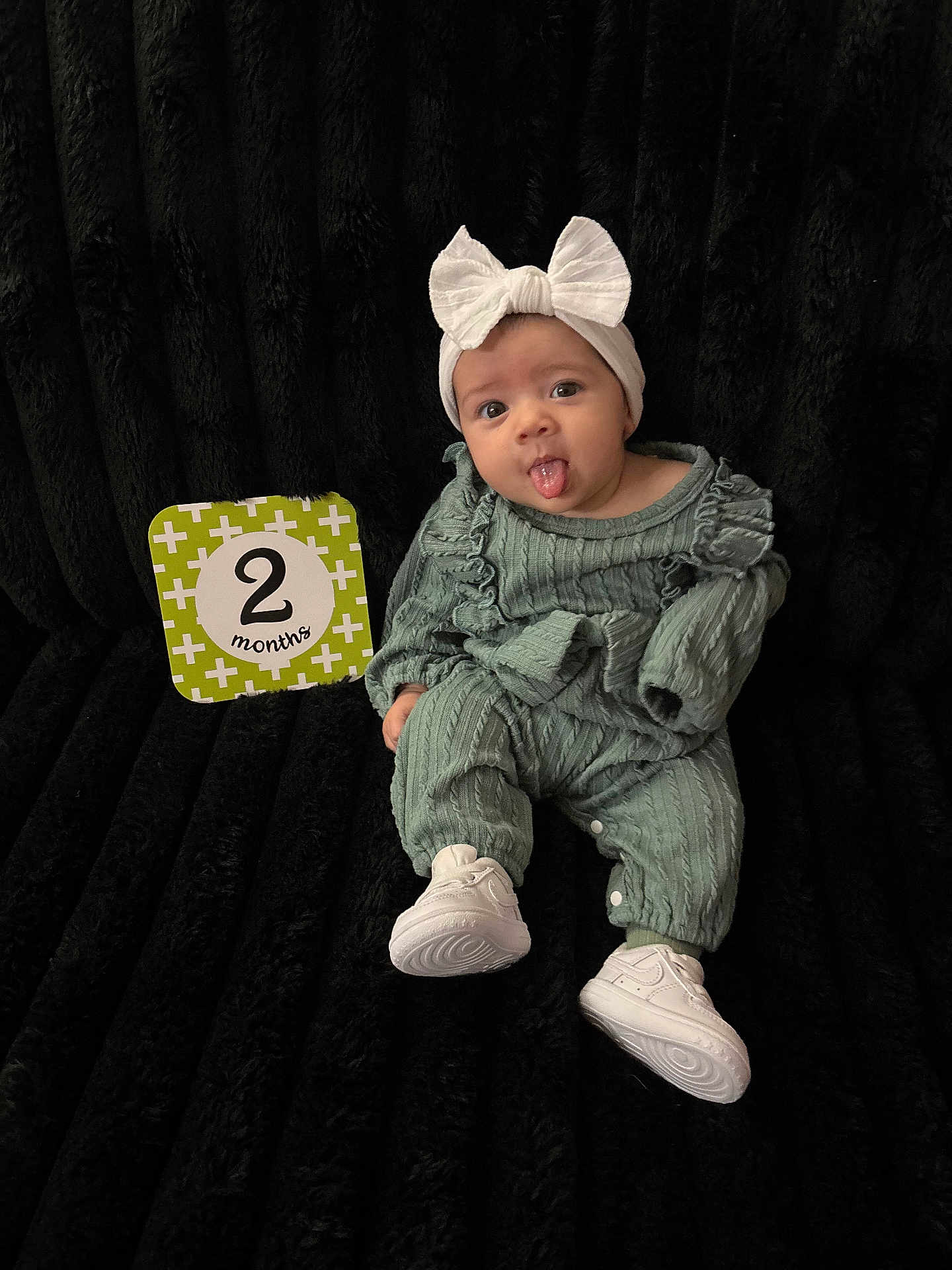 Azarah is registered to the contest to win money with this photo: 2_months, baby, black_background, child, clothing, cute, footwear, green_outfit, headband, infant, person, playful, portrait, sign, sitting, smile, sneakers, studio, tongue_out, white_bow