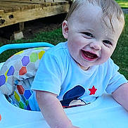 Everett is registered to the contest to win money with this photo: baby, child, high_chair, smiling, outdoor, grass, wooden_deck, greenery, toddler, happy, person, short_hair, blue_shirt, playful, sunlight, summer, daylight, hand, face, seat