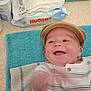 baby, infant, smiling, cap, striped_clothing, towel, diapers, baby_wipes, blurred_hand, happy, lying_down, indoor, baby_care_products, blue_towel, beige_towel, baby_hat, closeup, cheerful, person, child