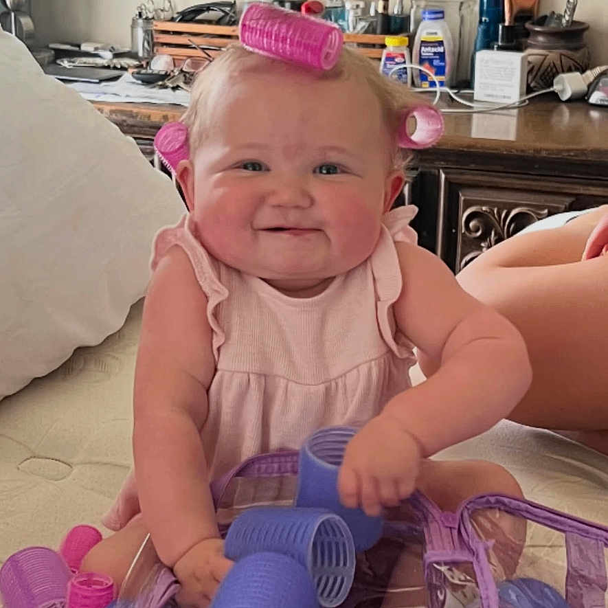 Lovelyn is registered to the contest to win money with this photo: baby, child, hair_rollers, pink, purple, smiling, indoors, bed, dresser, toiletries, cute, playing, person, infant, happy, skin, furniture, room, accessories, hair_care