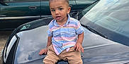 Nehemiah is registered to the contest to win money with this photo: toddler, child, car, black_car, hood, shoes, sneakers, pants, shirt, outdoor, daylight, suburb, vehicle, parked_car, curious, person, tree, grass, street, cloudy_sky