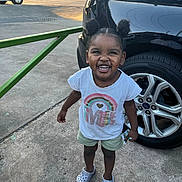 Naleighna is registered to the contest to win money with this photo: toddler, child, smile, hair_buns, tshirt, shorts, crocs, car, tire, concrete, outdoor, daylight, happy, person, standing, joyful, vehicle, sidewalk, casual_clothing, sunlight