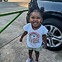 toddler, child, smile, hair_buns, tshirt, shorts, crocs, car, tire, concrete, outdoor, daylight, happy, person, standing, joyful, vehicle, sidewalk, casual_clothing, sunlight